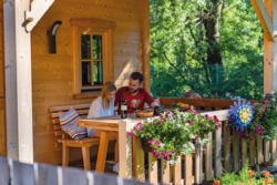 Zwei Personen sitzen auf der Veranda eines Holzhauses, trinken Wein und sehen sich eine Zeitschrift an. Im Vordergrund Blumen.